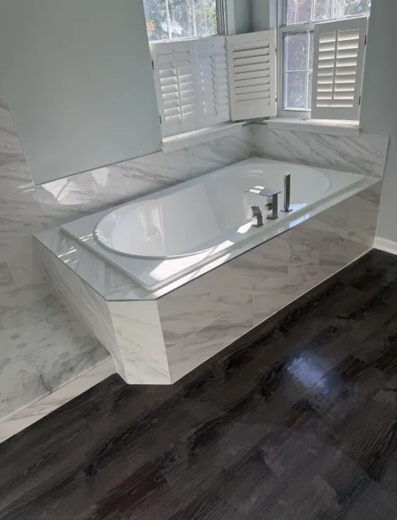 White tub set into a gray-veined tiled deck beneath shuttered windows, beside dark plank flooring.